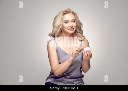Reife Erwachsene blonde Frau trägt Feuchtigkeitscreme auf, während sie eine Dose kosmetisches Mittel in der Hand hält, während sie die Kamera auf einem grau isoliert betrachtet. Stockfoto