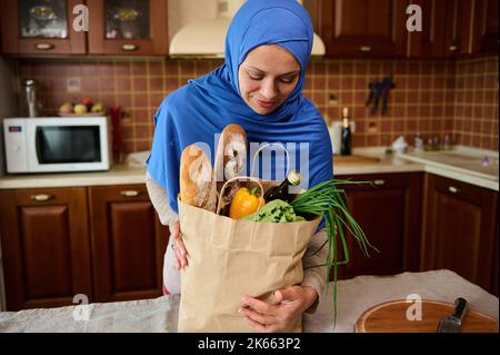 Die Frau aus dem Nahen Osten, die sich in einem Hijab befindet, kommt nach Hause und packt nach dem Einkaufen ein Päckchen mit Bio-Lebensmitteln aus Stockfoto