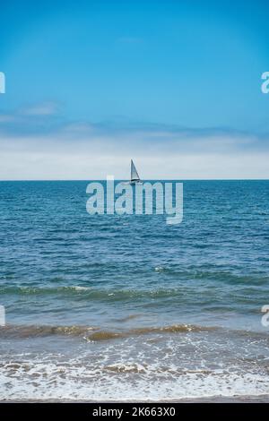 Kleines weißes Segelboot mit Blick auf den Ozean vom Butterfly Beach Santa Barbara California. Blauer monochromer vertikaler Hintergrund im Hochformat Stockfoto