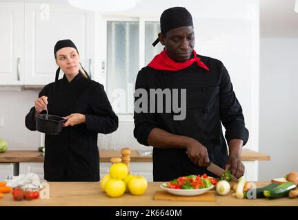 Fokussierter Koch in schwarzer Uniform Kochen in privater Küche mit weiblicher Kollegin Stockfoto