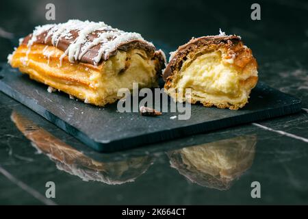 Hausgemachtes Eclair mit Mango- und Kokosnuss-Schlagsahne. In Milchschokolade getaucht. Stockfoto