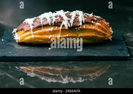 Hausgemachtes Eclair mit Mango- und Kokosnuss-Schlagsahne. In Milchschokolade getaucht. Stockfoto