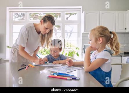 Kaukasische Mutter hilft ihren kleinen Kindern, ein Buch mit Farbstiften in der Küche zu Hause zu zeichnen Stockfoto