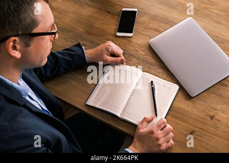 Junger seriöser Geschäftsmann schreibt etwas im Notebook, das mit dem liegenden Smartphone am Tisch sitzt. Mann trägt Brille füllt Notebook mit Stift, organisieren Stockfoto