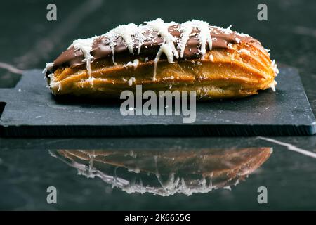 Hausgemachtes Eclair mit Mango- und Kokosnuss-Schlagsahne. In Milchschokolade getaucht. Stockfoto