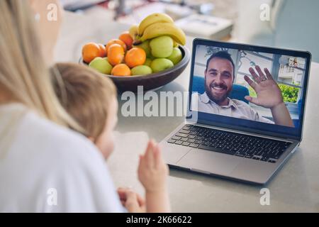 Mutter und Sohn sprechen zu Hause mit ihrem Mann über einen Videoanruf auf einem Laptop. Der kaukasische Vater aus großer Entfernung winkt seinem Sohn während einer Videokonferenz zu Stockfoto