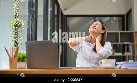 Erschöpfte junge asiatische Geschäftsfrau oder Büroangestellte, die an Nackenschmerzen oder Bürosyndrom leidet. Überfordert, schmerzhaft, Schulterschmerzen oder Fi Stockfoto