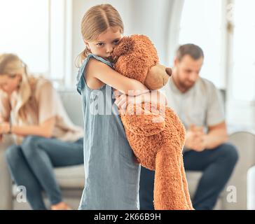 Ein verärgertes kleines Mädchen quetscht ihren Teddybär, während es traurig und deprimiert aussieht, während ihre Eltern im Hintergrund streiten. Denken Sie an ihre Eltern Stockfoto