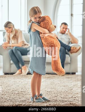 Ein verärgertes kleines Mädchen quetscht ihren Teddybär, während es traurig und deprimiert aussieht, während ihre Eltern im Hintergrund streiten. Denken Sie an ihre Eltern Stockfoto