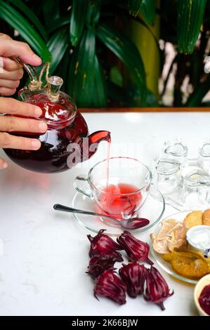 Hände halten Glas Teekannen mit Hibiskustee mit Tassen und kristallisierten Früchten und Marmelade auf einem Tisch Stockfoto