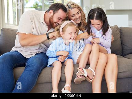 Verspielte kaukasische Eltern kitzeln ihre Kinder, während sie sich zu Hause auf einem Sofa entspannen. Sorglose kleine Mädchen verbringen Zeit mit liebevollen Eltern Stockfoto