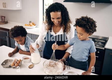 Glückliche afroamerikanische Mutter beim Backen mit ihren Söhnen. Zwei Brüder backen mit ihren Eltern in der Küche zu Hause. Kleiner Junge, der eine Schüssel mit Teig stischt Stockfoto