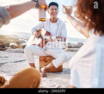 Nahaufnahme eines jungen asiatischen Mannes, der draußen am Strand für seine Freunde Gitarre spielt, während er Alkohol trinkt Stockfoto