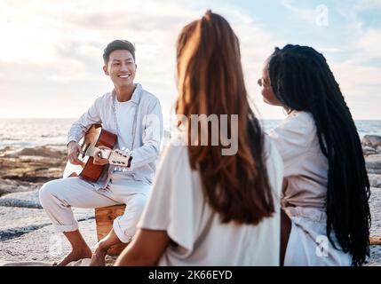 Ein junger Mann am Strand, der mit seinen beiden Freundinnen Gitarre spielt. Eine vielfältige Gruppe von Freunden, die an einem sonnigen Tag Musik hören Stockfoto