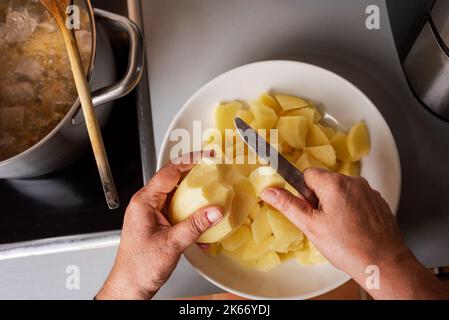 Hände der Frau schneiden Kartoffeln in Stücke in der Küche zu Hause, um den Topf mit dem geschmorten Fleisch hinzuzufügen.Nahaufnahme. Stockfoto