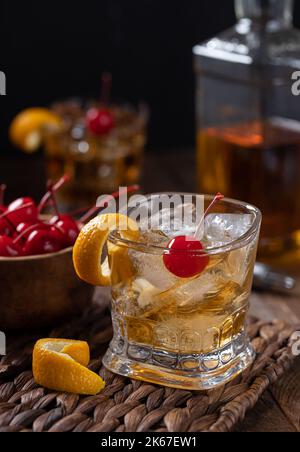 Altmodischer Cocktail mit Orangenschale und Kirsche auf einem Holztisch mit einer Schüssel mit Kirschen und einer Whiskey-Flasche Stockfoto
