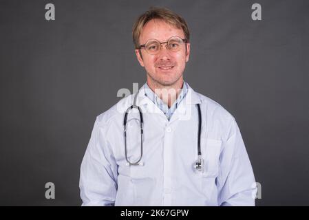 Glücklicher männlicher Arzt mit Stethoskop über dem Hals im medizinischen Mantel vor grauem Hintergrund Stockfoto