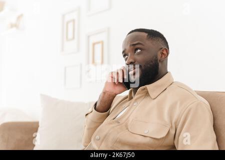 Schwarzer aufgeregt Kerl ist mit Smartphone, im Gespräch mit einem Freund, während auf dem Sofa zu Hause sitzen. Mobile Verbindung Stockfoto