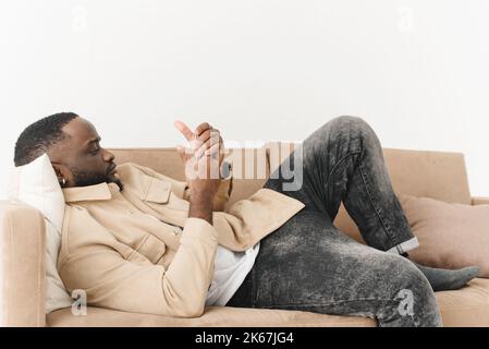 Junger afroamerikanischer Mann liegt auf dem Sofa und telefoniert. Stockfoto