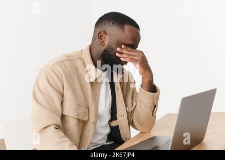 Müde depressive afroamerikanische Freiberuflerin, frustriert über Geschäftsversagen, Arbeit am Laptop, sich erschöpft von Kopfschmerzen, verärgert zu Hause. Stockfoto