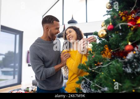 Bild eines glücklichen, vielfältigen Paares, das zu Hause weihnachten feiert und den Baum dekoriert Stockfoto