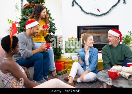 Bild von fröhlichen, vielfältigen Freunden, die zu Hause weihnachten feiern und sich unterhalten und heiße Schokolade trinken Stockfoto