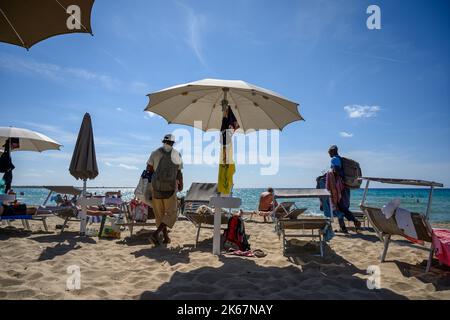Zwei Strandverkäufer, die gegen Ende der Saison unter Sonnenanbetern ihre Waren am Strand von Sottovento anbieten. Gallipoli, Apulien (Apulien), Italien. Stockfoto