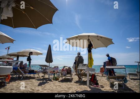 Zwei Strandverkäufer, die gegen Ende der Saison unter Sonnenanbetern ihre Waren am Strand von Sottovento anbieten. Gallipoli, Apulien (Apulien), Italien. Stockfoto