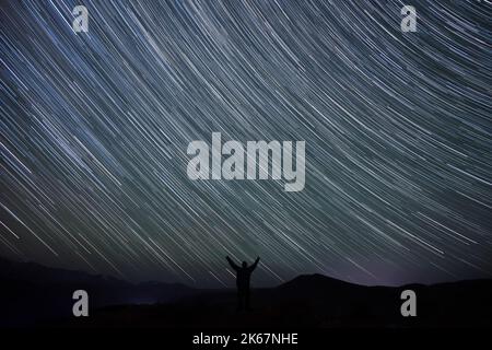 Abstrakte Nachtlandschaft mit Sternenhimmel mit Spuren von Sternen in Form von Spuren über den Bergen und der Kontur eines Mannes auf der Spitze des Mo Stockfoto