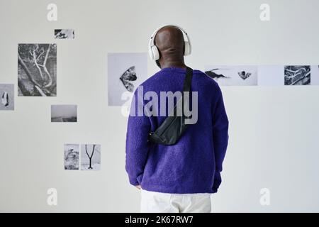 Rückansicht eines afrikanischen jungen Mannes in kabellosen Kopfhörern, der Führer hört und Fotos an der Wand ansieht Stockfoto