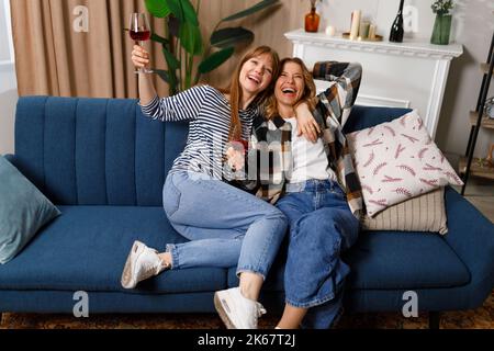 Fröhliche, reife Mutter und Erwachsene Tochter reden, lachen und umarmen, während sie zu Hause auf dem Sofa Wein trinken Stockfoto