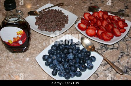 Obst- und Schokoladenchips und Sirup zum Beladen von Pfannkuchen und Waffeln auf kleinen weißen Tellern mit einem Löffel. Stockfoto