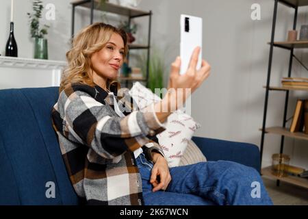 Attraktive Frau posiert vor der Smartphone-Kamera, während sie auf dem Sofa in der Wohnung sitzt Stockfoto