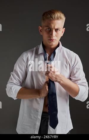 Porträt eines hübschen jungen Mannes, der in einem weißen Hemd posiert und auf grauem Hintergrund eine Krawatte gebunden hat. Klassische Männermode Stockfoto
