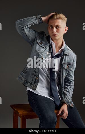 Porträt eines jungen Mannes mit blonden Haaren in Jeans Jacke isoliert auf grauem Hintergrund posiert. Lässiger Style. Brutaler Look Stockfoto