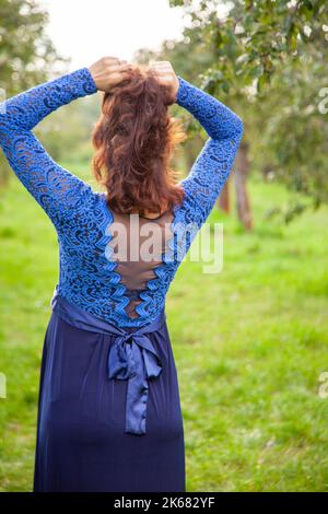 Eine Frau mit roten Haaren steht mit ihrem Rücken in einem Garten. Blick von hinten. Eine braunhaarige Frau in einem blauen Kleid geht spazieren. Ruhige Stimmung und Rückzug in der Natur. Stockfoto