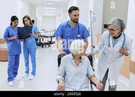 Freundlicher Arzt und Krankenschwester kümmern sich um die Gesundheit der älteren Frau, die auf medizinischen Rollstuhl mit intravenösen Tropfer in der Hand sitzt. Patientenversorgung bei Hosp Stockfoto