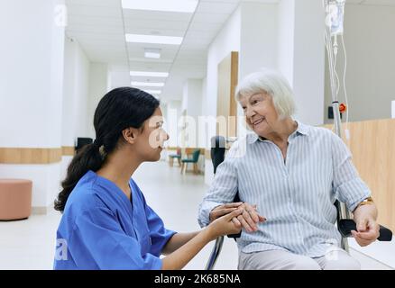 Psychologische Unterstützung von Krankenschwester zu älterer Frau, die im Rollstuhl auf dem Flur der medizinischen Klinik sitzt. Pflege von Menschen mit Behinderungen im Krankenhaus Stockfoto