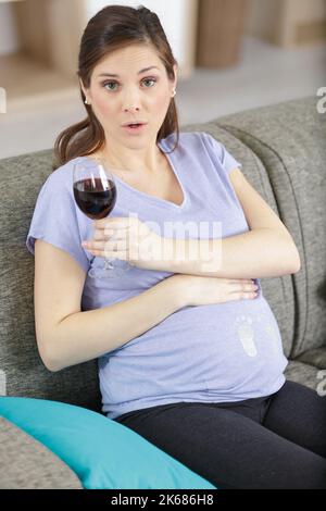 Eine Schwangerin trinkt Rotwein und sitzt auf dem Sofa Stockfoto