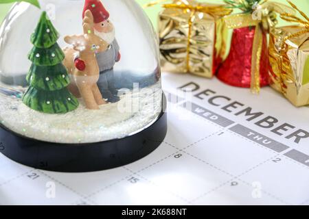 Countdown bis Weihnachten, festliche Schneekugel im Dezember-Kalender, Weihnachtsdekorationen wie Geschenke und Glocken im Hintergrund Stockfoto