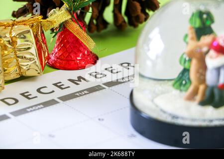 Weihnachtszeit, Dezemberkalender umgeben von Weihnachtsdekorationen, goldumhüllten Geschenken, glänzenden roten Glocken, Kieferkegel, Schneekugel außerhalb des Fokus im Vordergrund Stockfoto