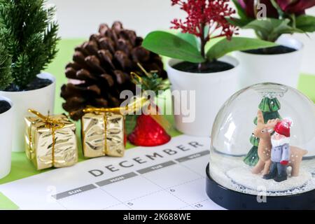 Weihnachtskonzept, festliche Schneekugel im Dezember-Kalender, außer Fokus im Hintergrund sind Dekorationen wie Pflanzen, Kieferkegel, Gold verpackte Geschenke, Stockfoto