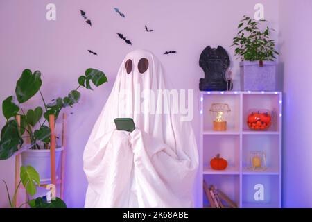 Der Halloween-Geist verwendet ein Mobiltelefon, um im Internet zu surfen, Online-Shops und Märkte zu durchsuchen. Der Geist macht an Halloween eine Bestellung von einem Smartphone. Neonlicht. Stockfoto