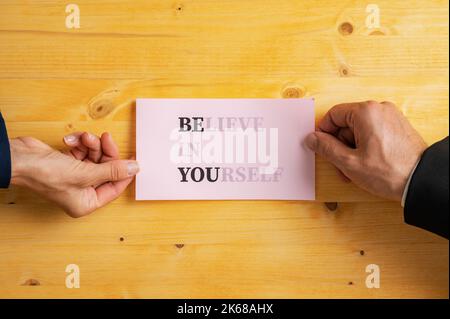 Männliche und weibliche Hände halten rosa Papier mit einem glauben an sich selbst Zeichen geschrieben, auf sie mit Worten WERDEN SIE mehr betont. Über gelbem Holzboden Stockfoto