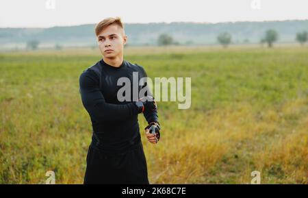 Ein junger Mann, der während der goldenen Stunde im Wald trainiert, den Puls streckt und misst, während er die Natur genießt Stockfoto