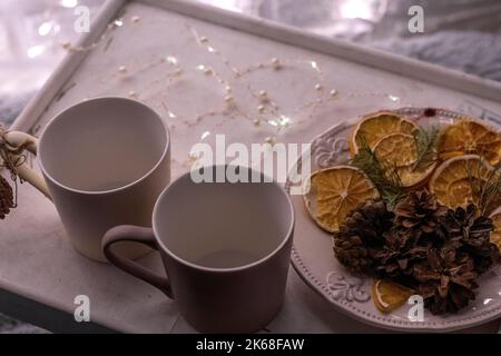 Zwei weiße Tassen auf einem Holztablett stehen auf einem Bett mit einer flauschigen Decke und einer Girlande im Hintergrund. Neujahrsstimmung Stockfoto