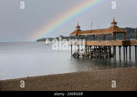 Der hölzerne Pier am Llanquihue-See unter einem düsteren Himmel mit Regenbogen in Frutillar, Chile Stockfoto