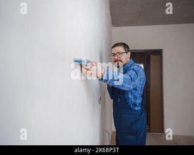 Junger Mann in blau Arbeitsanzug tun Reparatur Wohnung. Renovierungskonzept für das Haus. Kurzer Schuss eines gutaussehenden bärtigen jungen Mannes, der während der Heimverbesserung ein Level-Tool hält Stockfoto