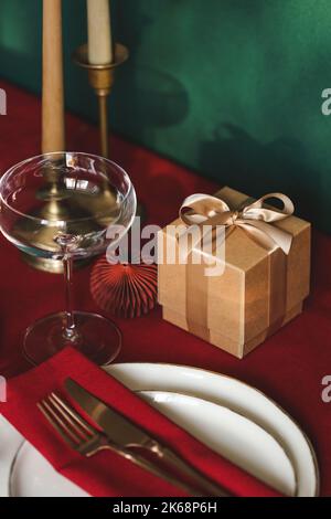 Weihnachtliche Tischeinstellung zwischen Geschenkbox und Kerzen auf roter Tischdecke. Konzept des Weihnachts- und Neujahrsdinners. Stockfoto