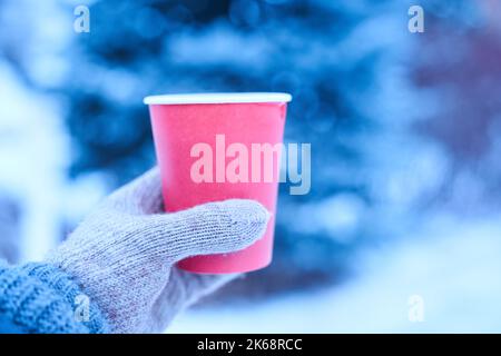 Becher in der Hand im Winter mit einem heißen Getränk, Modell für Ihren Text, Winterspaziergang mit heißem Tee in Handschuhen, roter Papierbecher eco auf blauem Hintergrund. Hochwertige Fotos Stockfoto